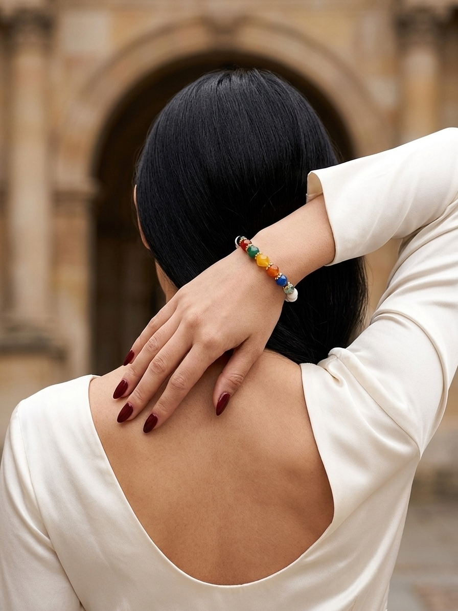 A woman viewed from behind wearing a cream-colored top, with her hand resting on her neck to display a colorful beaded bracelet. The background is a softly blurred stone archway.