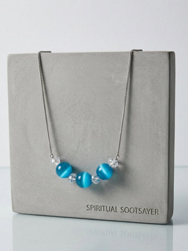A silver chain necklace with three bright blue cat-eye beads and small clear crystal accents, displayed draped over a gray square card labeled 'SPIRITUAL SOOTSAYER'.