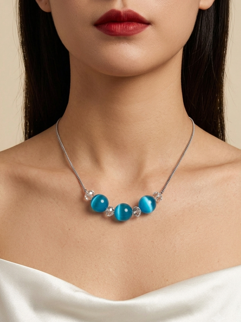 A close-up of a model wearing a silver necklace with four round turquoise-blue beads and small clear crystal accents, paired with a white off-the-shoulder top against a warm beige background.