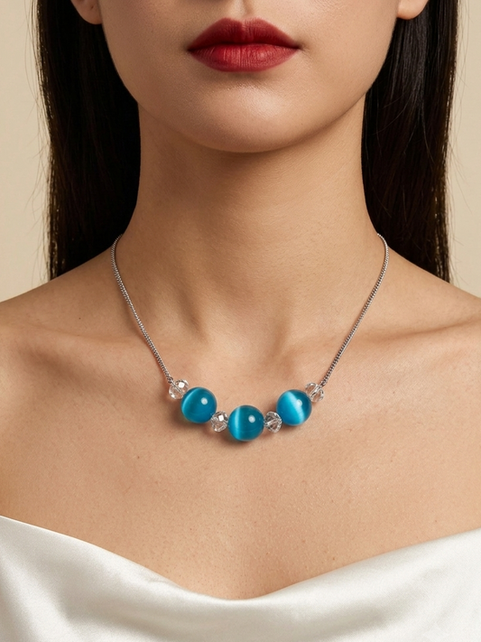 A close-up of a model wearing a silver necklace with four round turquoise-blue beads and small clear crystal accents, paired with a white off-the-shoulder top against a warm beige background.