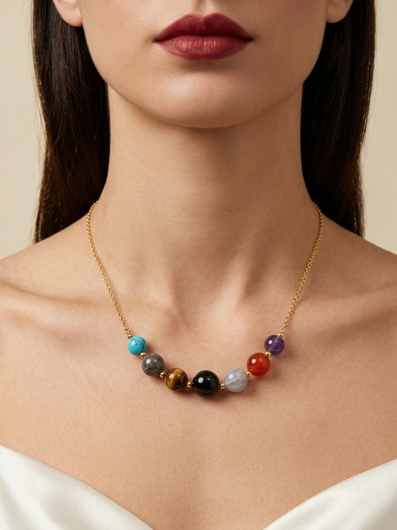 A close-up of a model wearing a delicate gold chain necklace with seven colorful round gemstone beads, styled with a white satin top against a warm beige background.