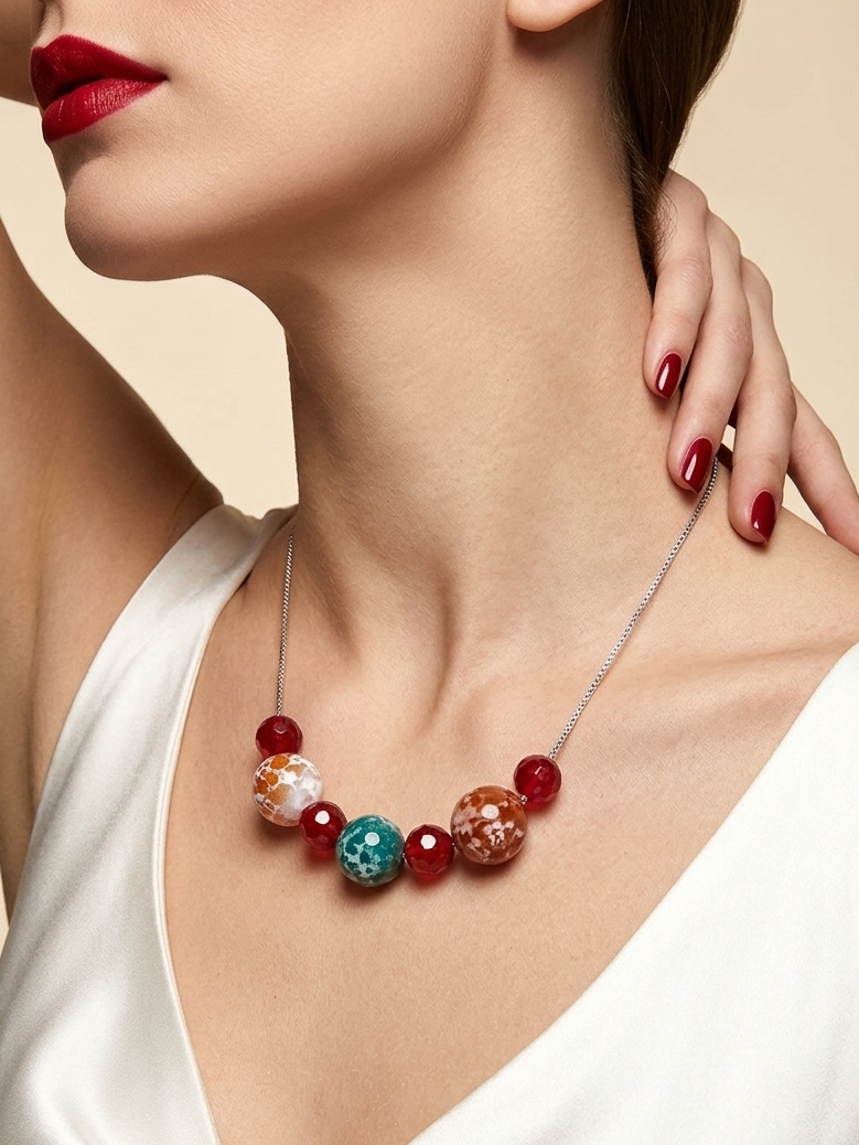A close-up of a model wearing a silver chain necklace with red beads and two decorative glass beads, styled with a white satin top against a beige background.