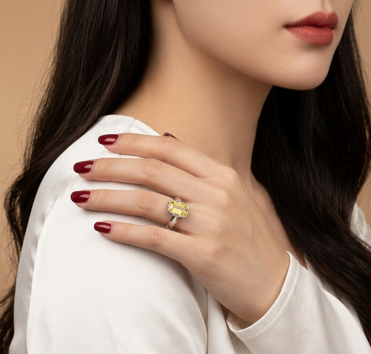 A close-up lifestyle shot of a woman wearing a yellow marquise-cut gemstone ring on her finger, resting her hand on her shoulder. She is wearing a white satin top against a warm beige background.