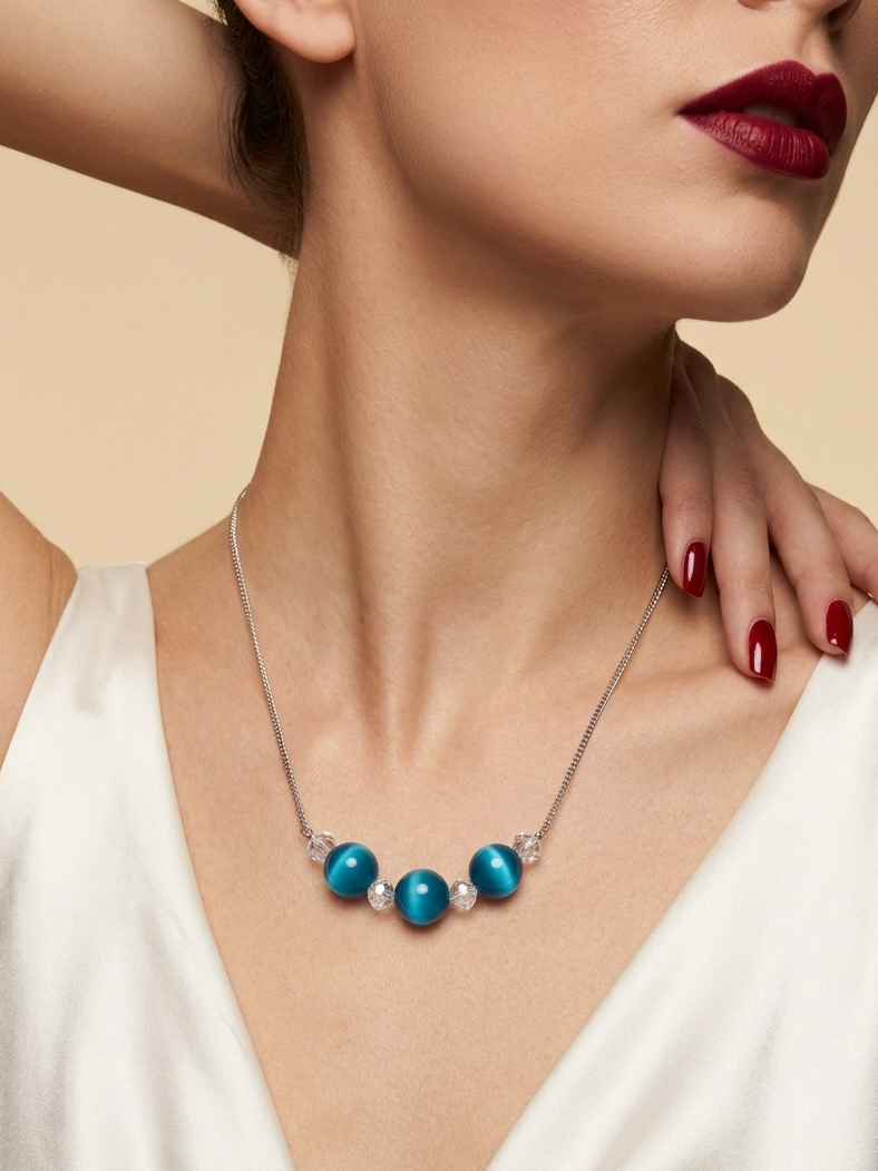 A close-up of a woman wearing a silver necklace with three round turquoise-blue beads and small clear stones, paired with a white satin top against a warm beige background.