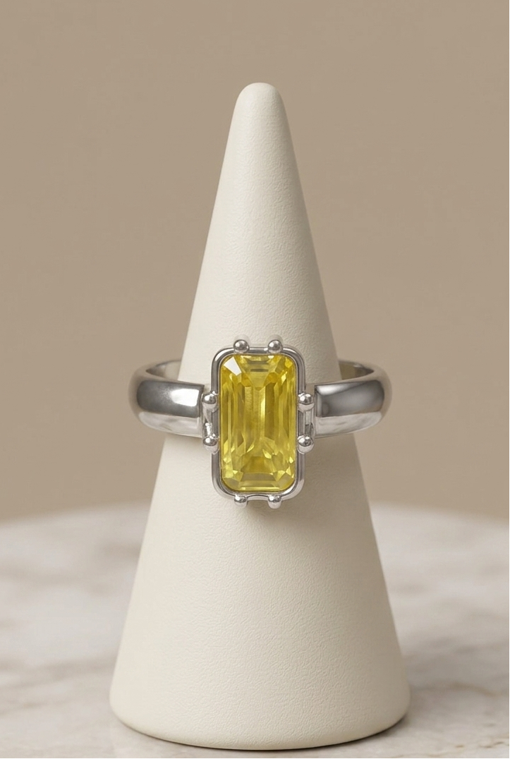 A silver ring with a rectangular yellow gemstone, displayed on a cream cone stand against a neutral beige background.