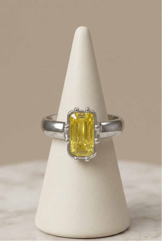 A silver ring with a rectangular yellow gemstone, displayed on a cream cone stand against a neutral beige background.
