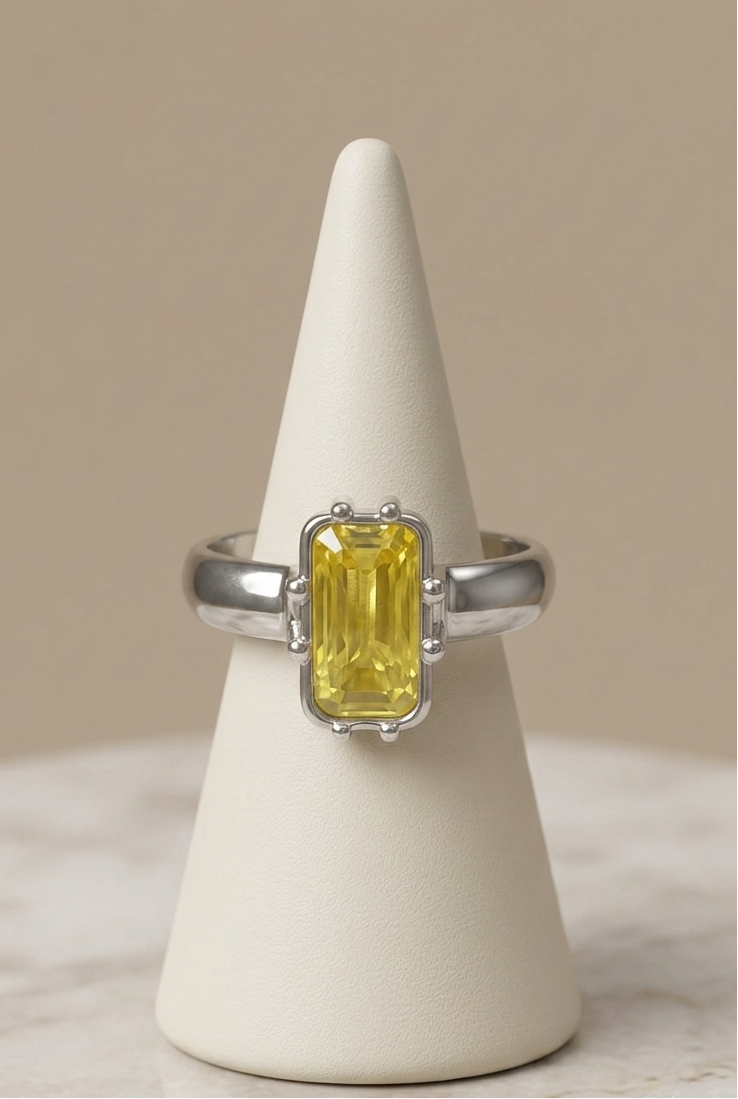 A silver ring with a rectangular yellow gemstone, displayed on a cream cone stand against a neutral beige background.