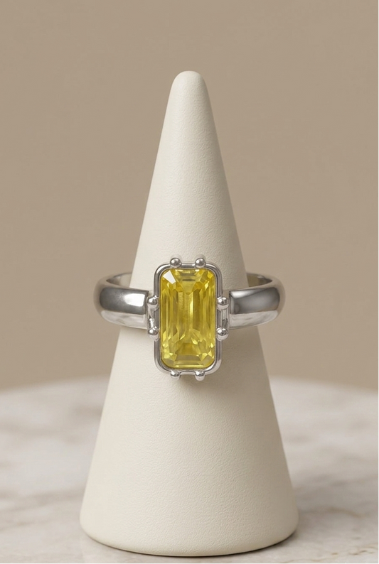 A silver ring with a rectangular yellow gemstone, displayed on a cream cone stand against a neutral beige background.