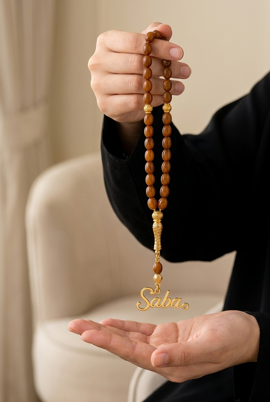 A person in a black long-sleeve top holds a strand of wooden prayer beads with a gold-tone metal tassel and a custom nameplate spelling 'Saba' in gold, set against a softly blurred indoor background.