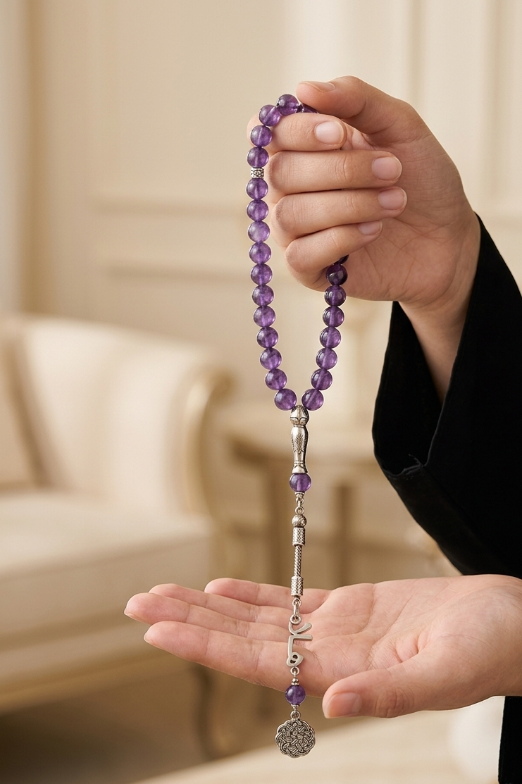 A person holds a purple beaded prayer rope in their hand, with a silver-toned tassel and small silver charm resting in their palm. The background shows a softly blurred indoor setting with warm beige tones.