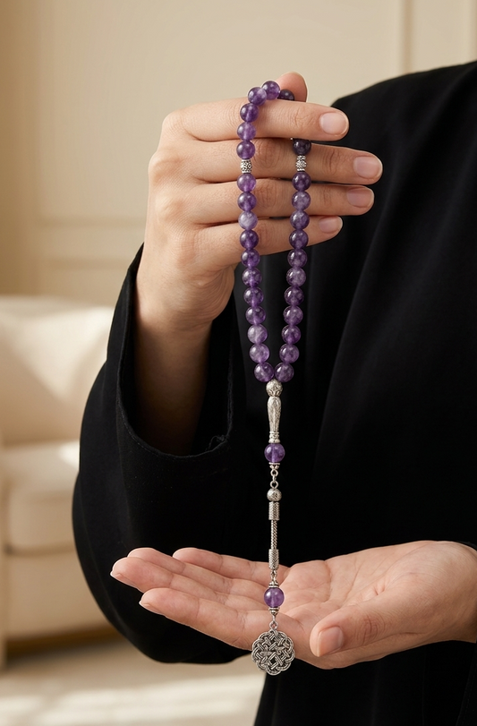 A person in a black robe holds up a strand of purple gemstone prayer beads with silver-toned metal accents and a round silver pendant in both hands, set against a softly blurred beige interior background.