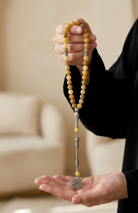 A person in a black robe holds a strand of yellow and cream beads with silver-toned spacer beads and a round silver pendant over their open palm, with a softly blurred indoor background.