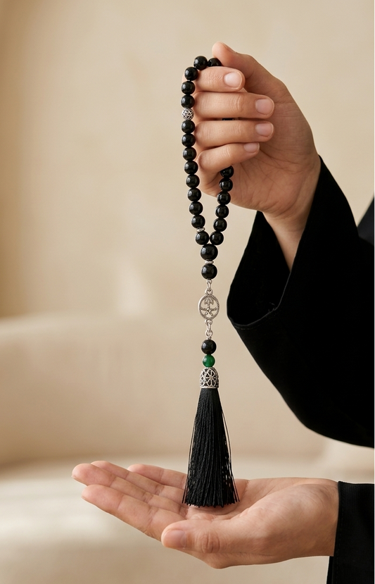 A person in a black robe holds a black beaded tassel necklace or prayer bead strand in both hands against a softly blurred beige indoor background.