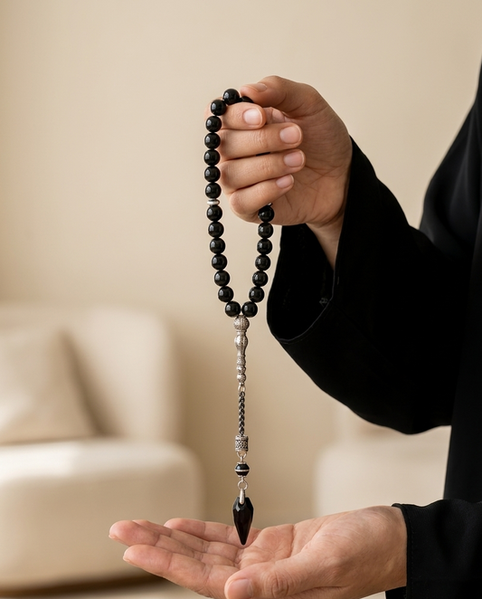 A person in a black robe holds a strand of black prayer beads (misbaha) over their open palm in a softly lit, modern indoor setting.
