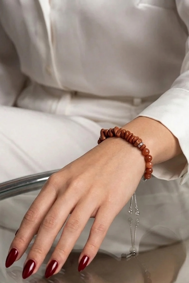 A close-up of a person's hand wearing a brown beaded bracelet with a silver chain tassel, while wearing a white button-up shirt.