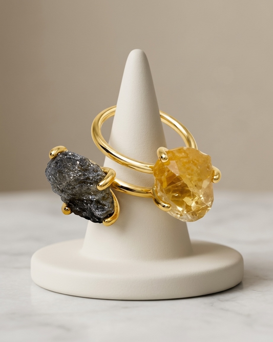Two gold rings displayed on a white cone ring stand. One ring features a rough dark gray stone, and the other features a faceted yellow stone, set against a neutral beige background.