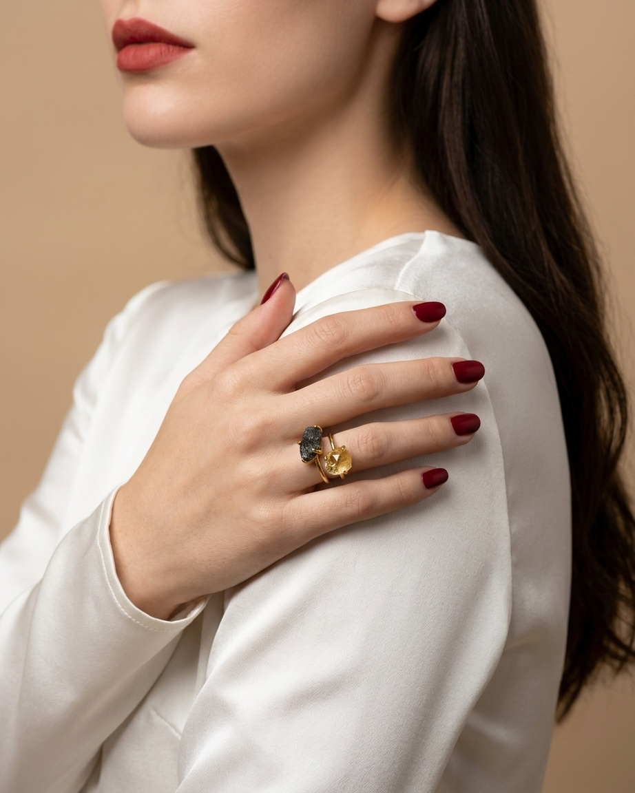 A close-up lifestyle shot of a woman wearing a gold ring with a dark blue stone and a small yellow gemstone, styled with a white satin top against a warm beige background.