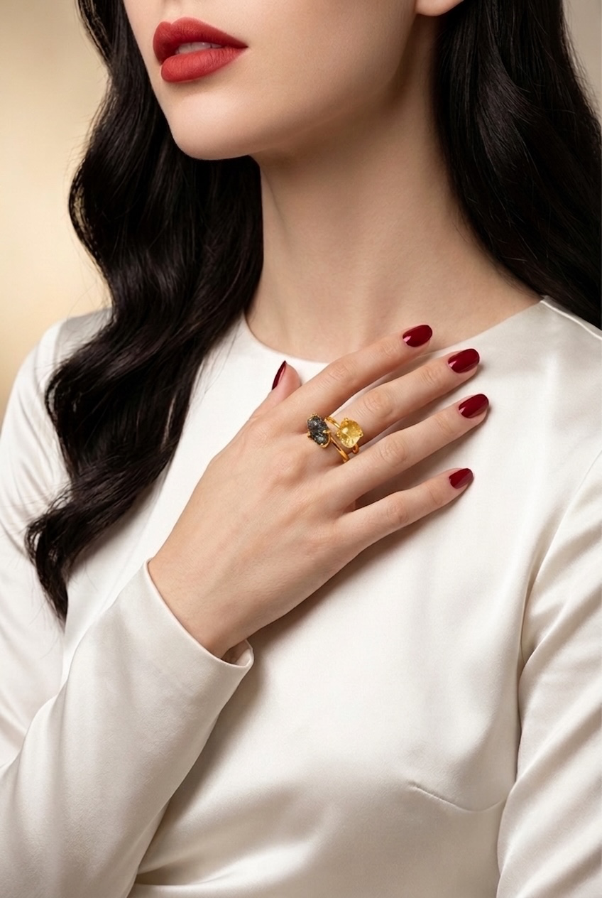 A close-up lifestyle shot of a woman wearing a gold ring with a dark gray marquise-cut stone and a yellow marquise-cut stone, styled with a white satin top and dark red lipstick.