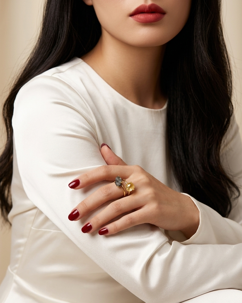 A close-up of a woman wearing a white satin long-sleeve dress, showcasing her hand with dark red manicured nails and two rings, one of which is a gold ring with a blue stone.