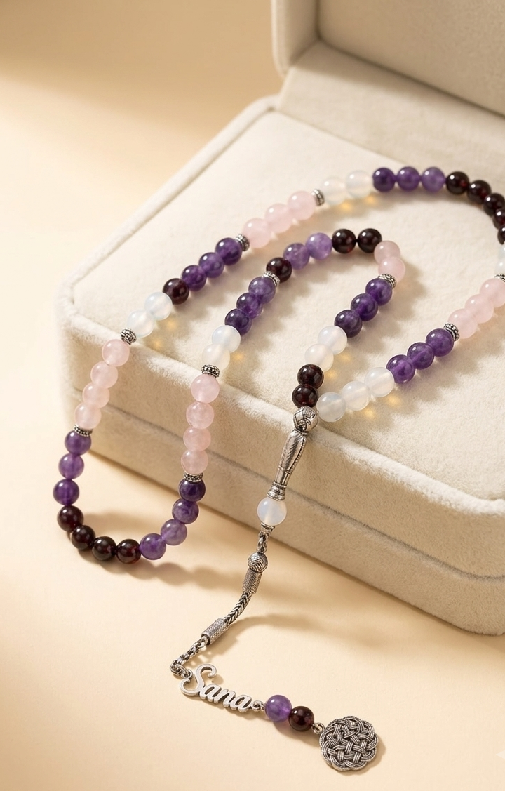 A beaded necklace with purple, white, and pink gemstone beads, a silver-toned chain, and a dangling script charm reading 'Sana' with a round textured pendant, displayed on a cream-colored jewelry box.
