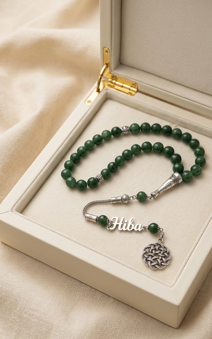A green beaded bracelet with a silver nameplate and circular pendant, displayed inside an open cream-colored jewelry box on a soft beige fabric background.