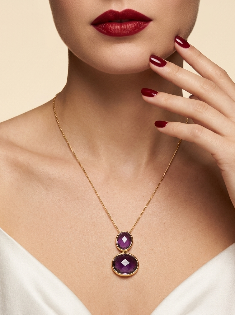 A close-up of a model wearing a delicate gold necklace with two stacked oval purple gemstones, paired with a white satin top and dark red lipstick.