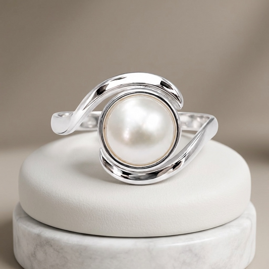 A polished silver ring featuring a large, round white pearl in the center, displayed on a round white cushion atop a marble stand against a soft beige background.