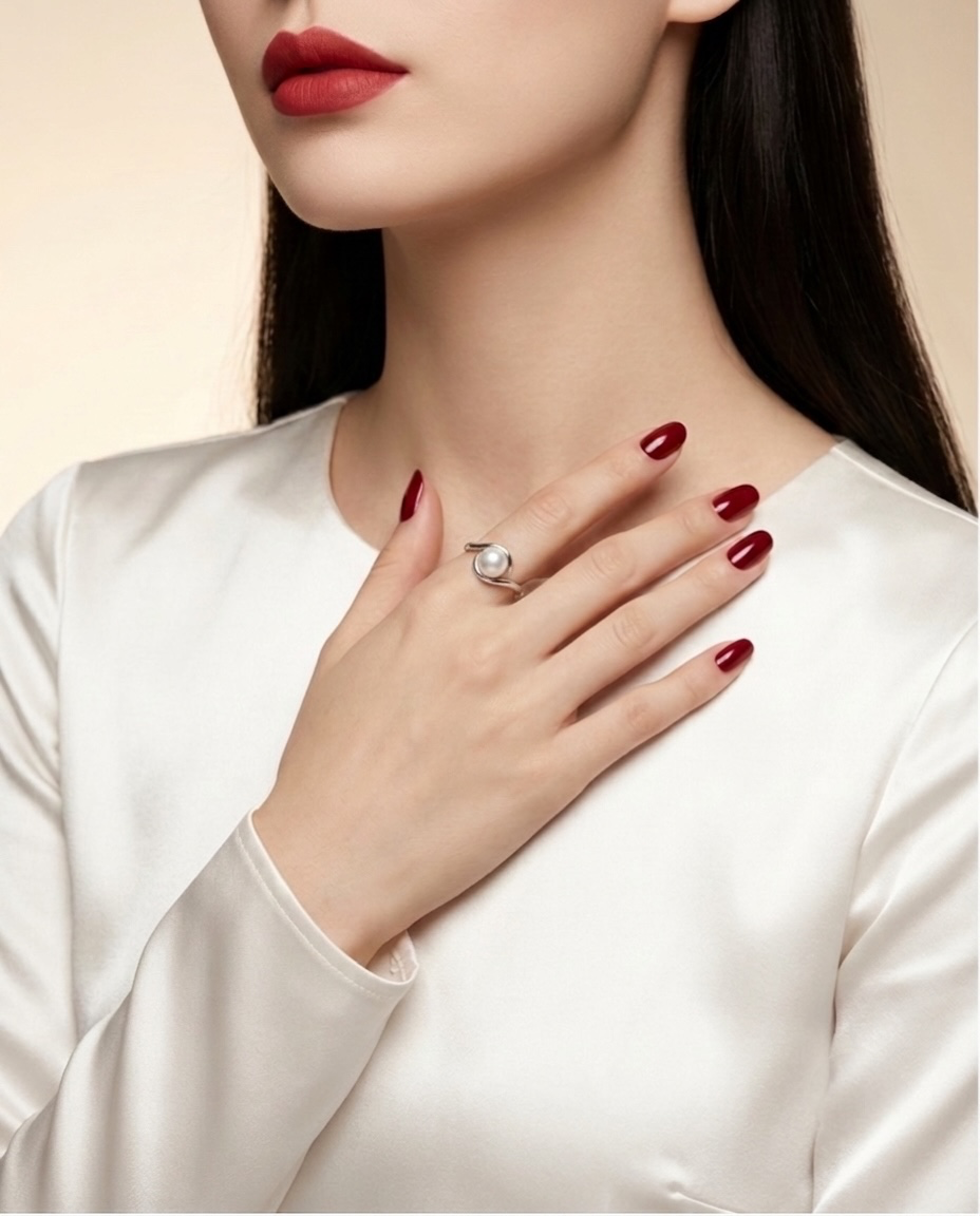 A close-up of a woman wearing a silver ring with a white stone on her finger, dressed in a white satin top against a warm beige background.