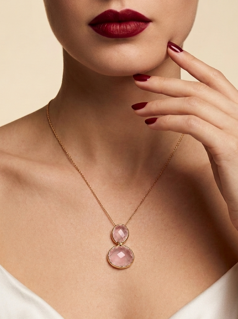 A close-up of a model wearing a delicate gold chain necklace with two stacked round pink gemstones, styled with a white satin top and dark red lipstick.