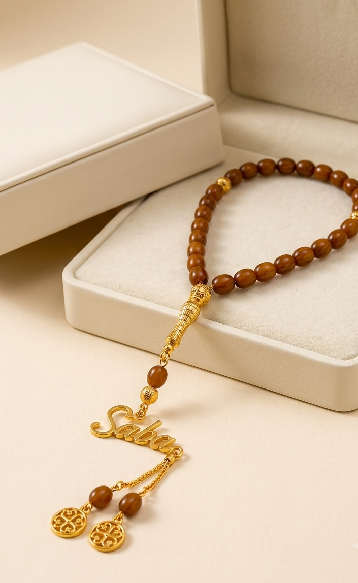 A beaded necklace with brown wooden beads and gold-toned metal accents, including a script nameplate charm and circular dangling charms, displayed on a cream-colored jewelry box.