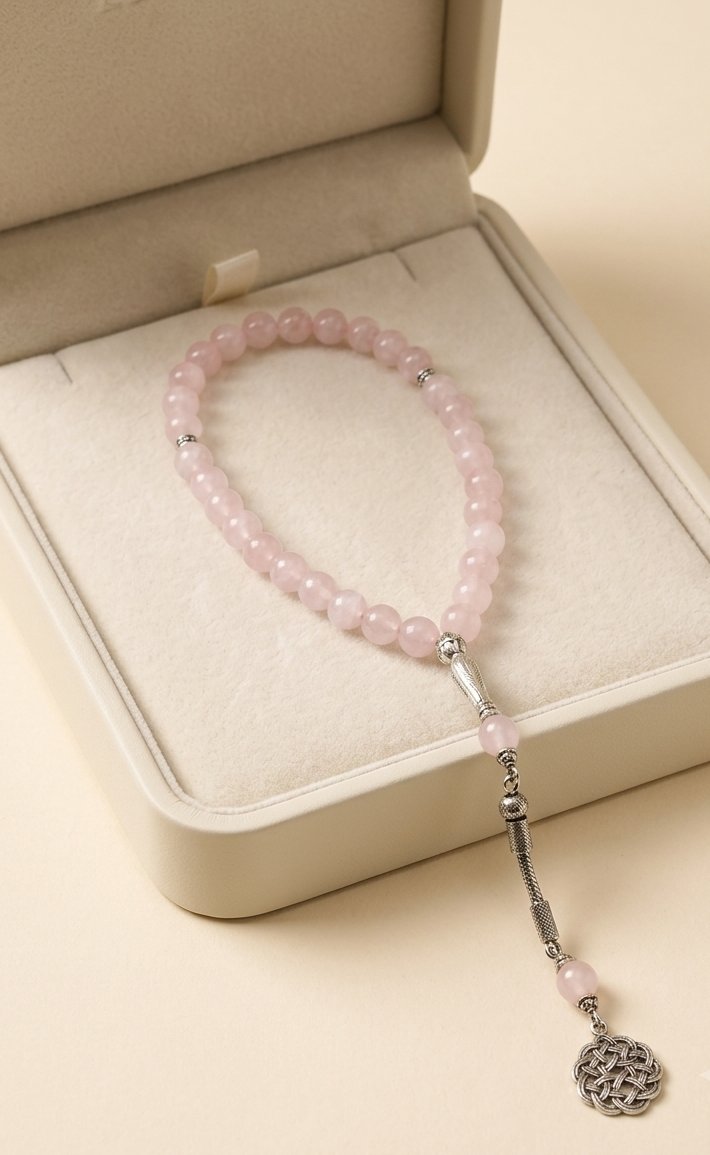 A pink beaded bracelet with silver-toned spacer beads and a dangling silver-toned tassel charm, displayed inside an open cream-colored jewelry box on a warm beige background.