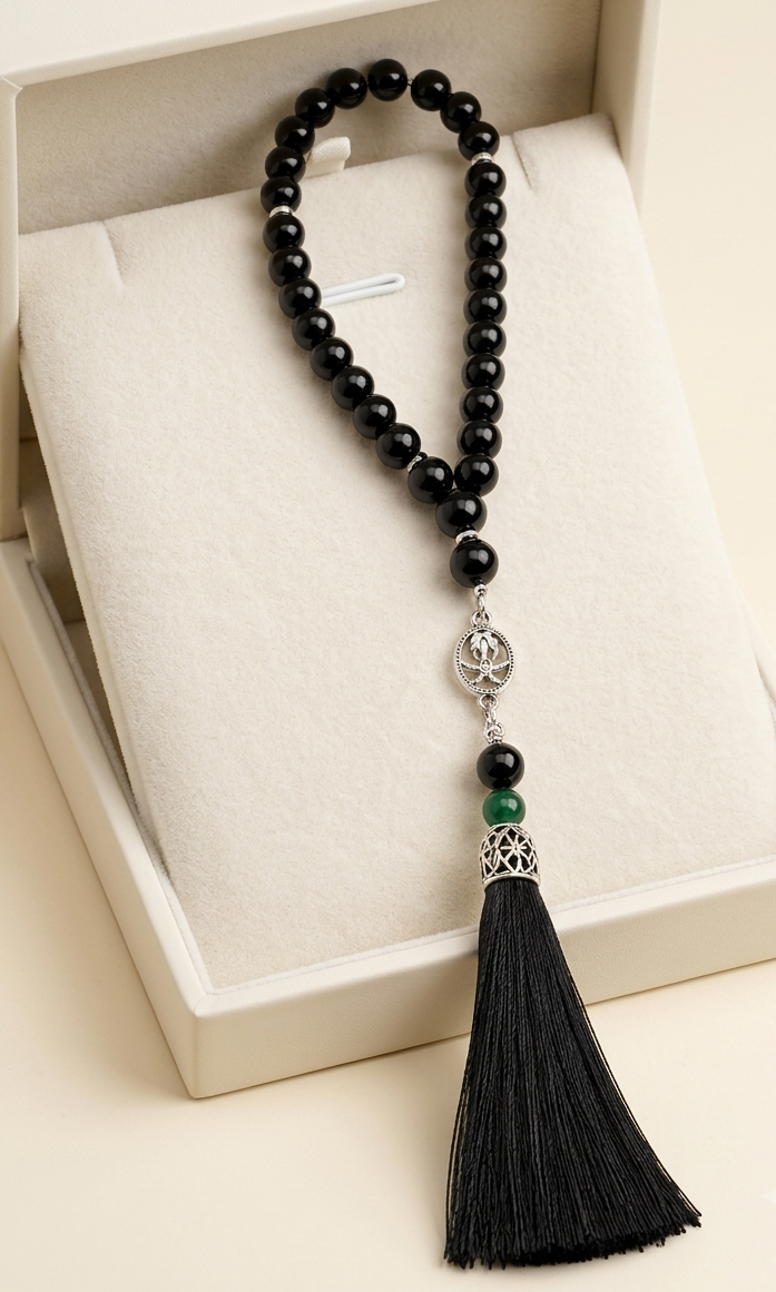 A black beaded tassel necklace with silver-toned metal accents and a small green bead, displayed inside an open cream-colored jewelry box.