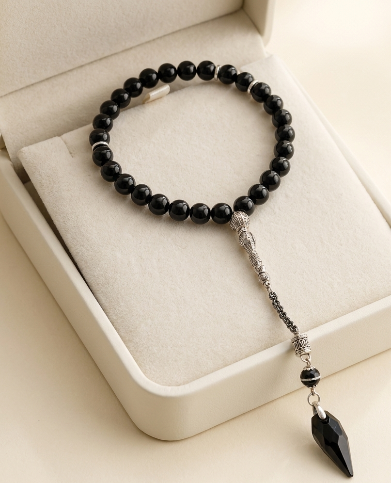 A black beaded bracelet with a silver-toned chain and a faceted black teardrop pendant, displayed inside an open cream-colored jewelry box.