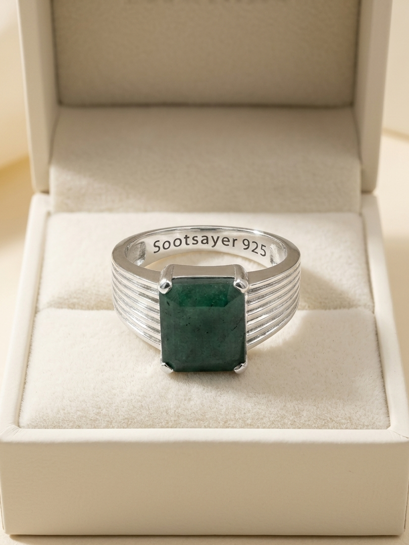 A silver ring with a rectangular green emerald-cut gemstone, displayed inside an open cream-colored jewelry box. The inside of the band is engraved with 'Sootsayer 925'.