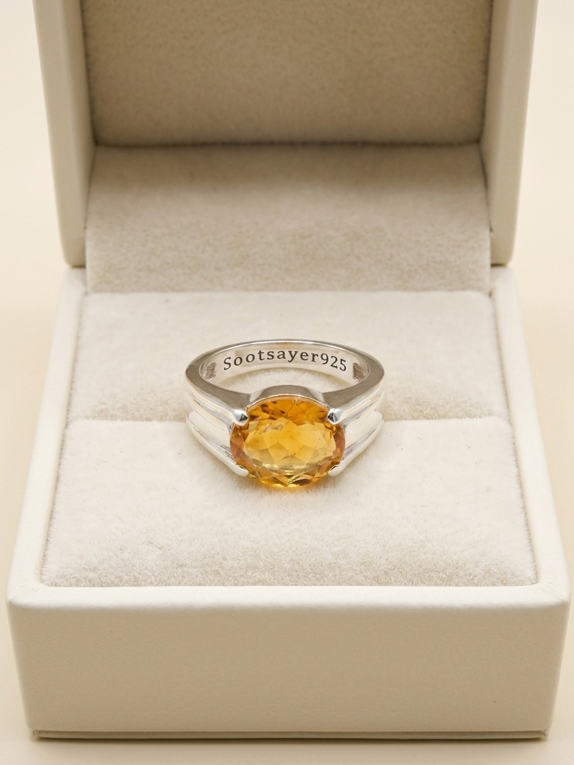 A silver ring with a round faceted yellow gemstone, displayed inside an open cream-colored jewelry box. The inner band of the ring is engraved with '580tsayer925'.