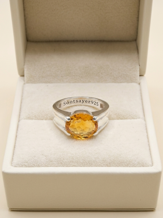 A silver ring with a round faceted yellow gemstone, displayed inside an open cream-colored jewelry box. The inner band of the ring is engraved with '580tsayer925'.