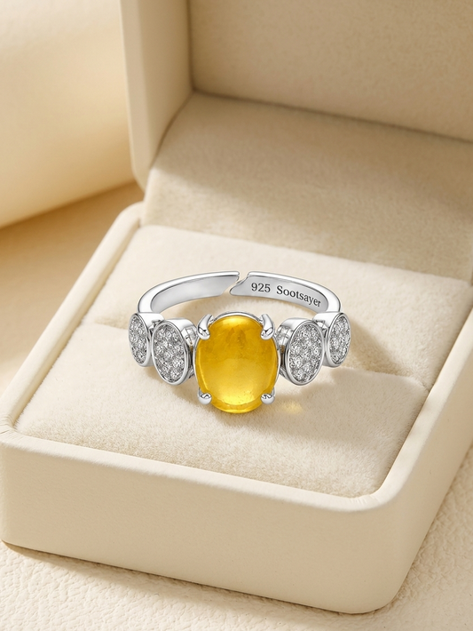 A silver-toned ring with a large oval yellow gemstone and smaller clear stones on either side, displayed in an open cream-colored jewelry box.