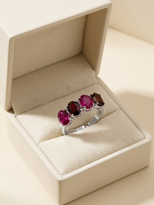 A silver ring with three oval-cut deep pinkish-red gemstones displayed in an open cream-colored jewelry box.