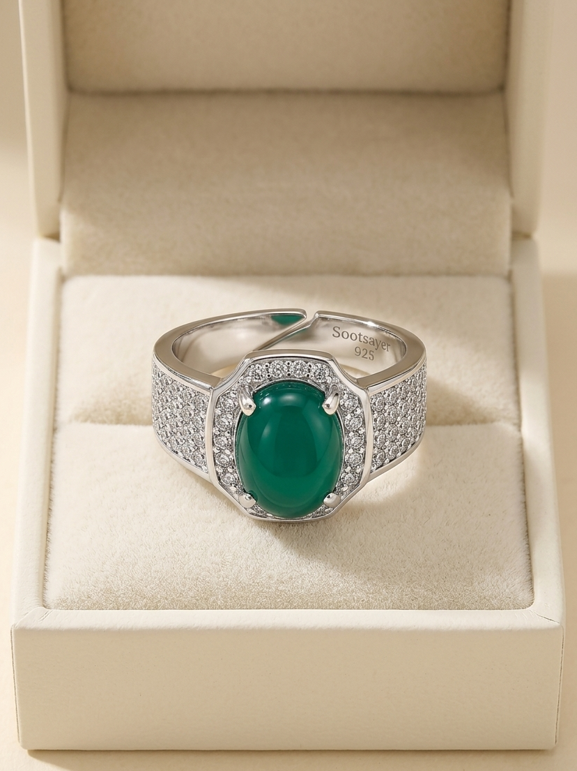 A silver ring with a large oval green cabochon gemstone and pavé-set side stones, displayed inside an open cream-colored jewelry box.