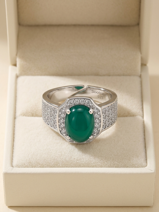 A silver ring with a large oval green cabochon gemstone and pavé-set side stones, displayed inside an open cream-colored jewelry box.