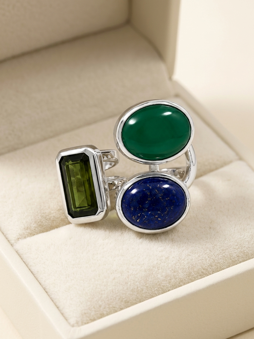 A silver ring with three gemstone settings displayed inside a cream-colored jewelry box. The ring features a large oval emerald-cut green stone, a smaller oval deep blue stone with gold flecks, and a rectangular green stone in a bezel setting.
