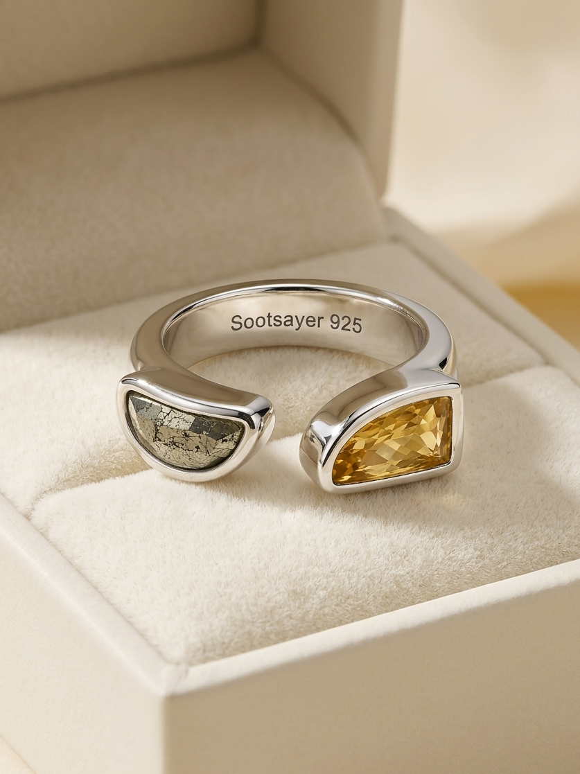 A silver open-bend ring displayed in a cream-colored jewelry box. The ring features a polished silver band stamped with 'Sootsayer 925' and two distinct stone inlays: a textured gray stone on one side and a faceted yellow-gold stone on the other.