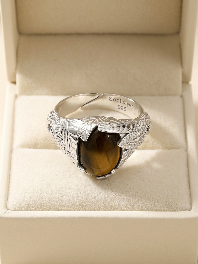 A sterling silver ring with a carved leaf design and a brown tiger's eye stone, displayed inside an open cream-colored jewelry box.
