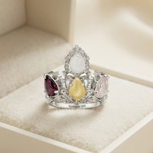 A silver crown-shaped ring with three teardrop gemstones (white, yellow, and burgundy) and small clear stones, displayed on a cream-colored jewelry box cushion. The band is engraved with 'Scottyner 925'.