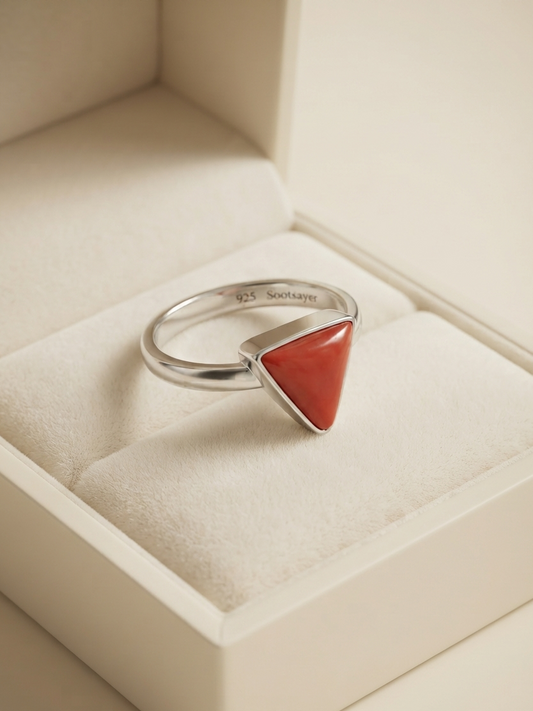 A silver ring with a triangular coral-red stone, displayed inside a cream-colored jewelry box.