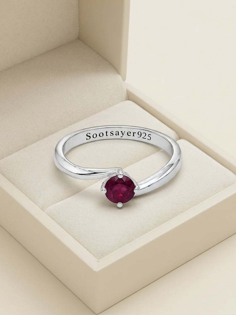 A sterling silver ring with a round deep red gemstone, displayed inside a cream-colored jewelry box. The inside of the band is engraved with 'Sootsayer925'.