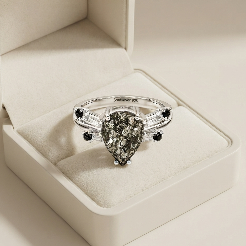A silver ring with a teardrop-shaped pyrite center stone and small black gemstone accents, displayed in an open cream-colored jewelry box.