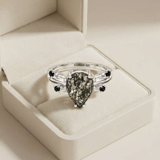 A silver ring with a teardrop-shaped pyrite center stone and small black gemstone accents, displayed in an open cream-colored jewelry box.
