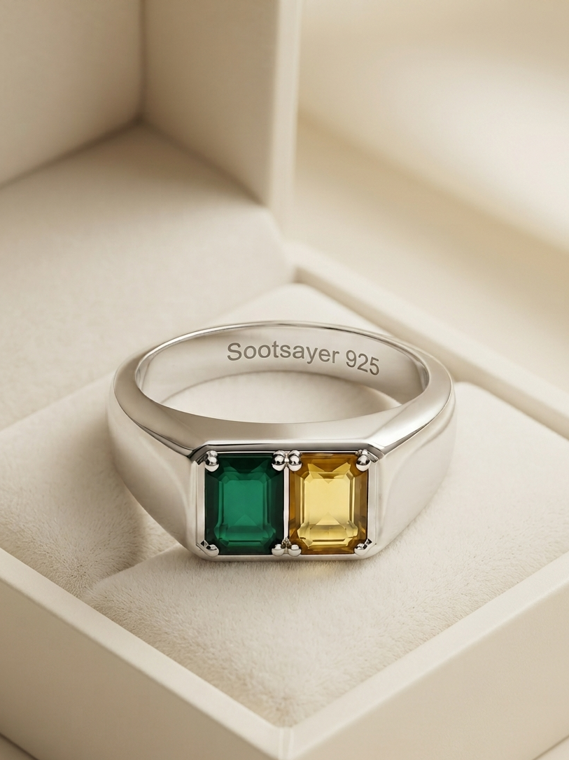 A silver 925 ring with two rectangular emerald-cut gemstones, one green and one yellow, displayed inside a cream-colored jewelry box. The inside of the band is engraved with 'Sootsayer 925'.