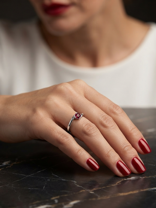 A close-up of a woman's hand resting on a dark marble surface, wearing a silver ring with a small red gemstone. The model has red-painted nails and is wearing a white top.
